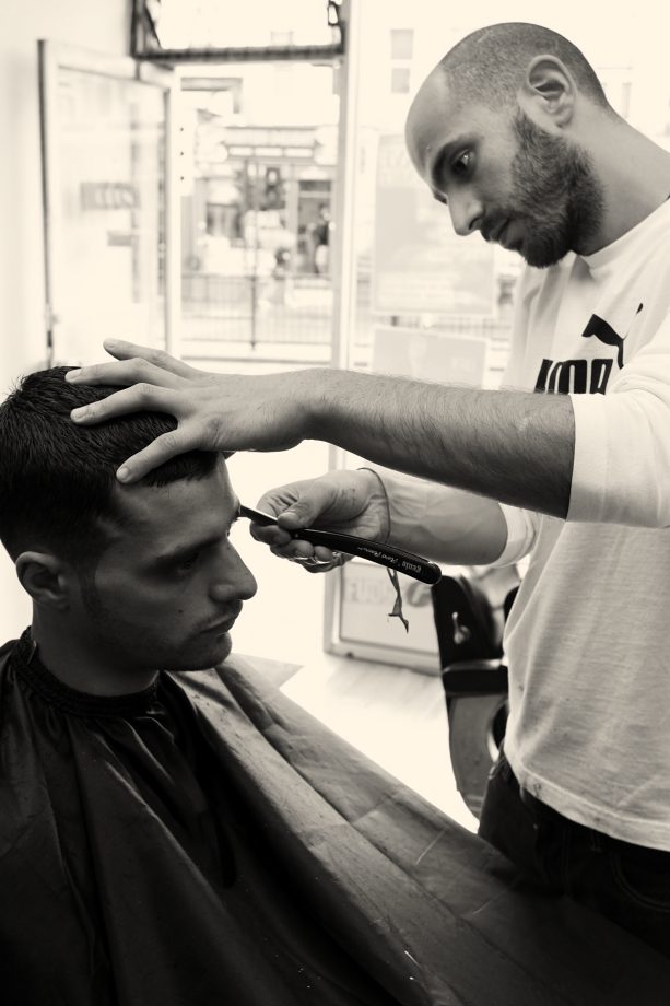 A barber tidying a hairline with a razor, Kentish Town Road through the window